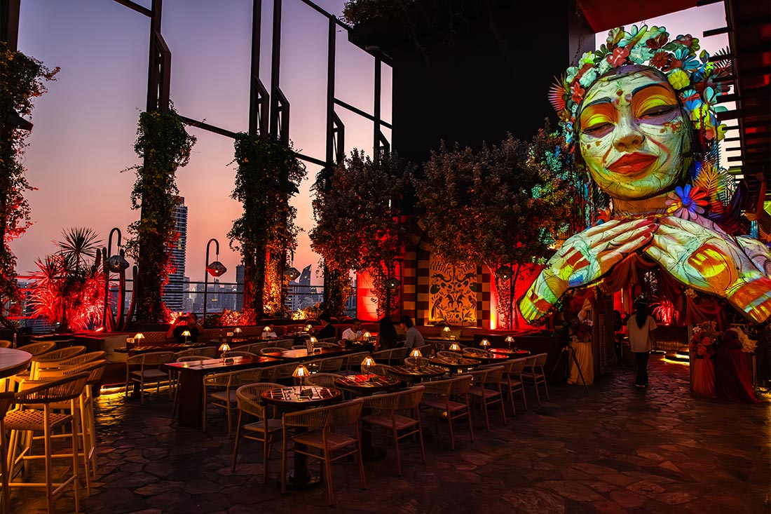 Stylish rooftop restaurant with warm red lighting and lush greenery surrounds modern dining tables at sunset. A massive colorful sculpture of a serene face decorated with flowers overlooks the space, creating a vibrant and artistic atmosphere against the evening skyline.
