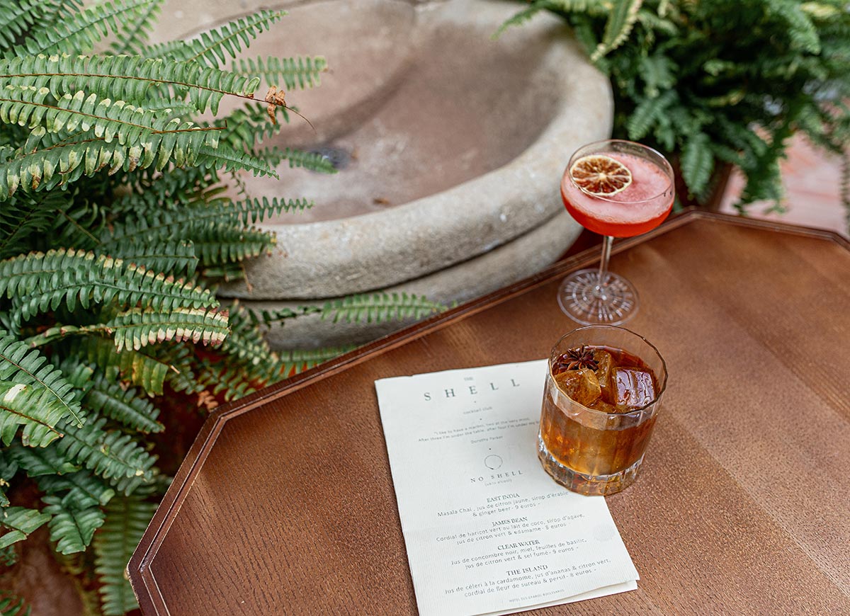 Two craft cocktails rest on a wooden table beside a menu in a lush indoor courtyard filled with ferns and greenery. One drink is served over ice with star anise garnish, while the other features a dried citrus slice, creating an elegant and inviting cocktail bar atmosphere.