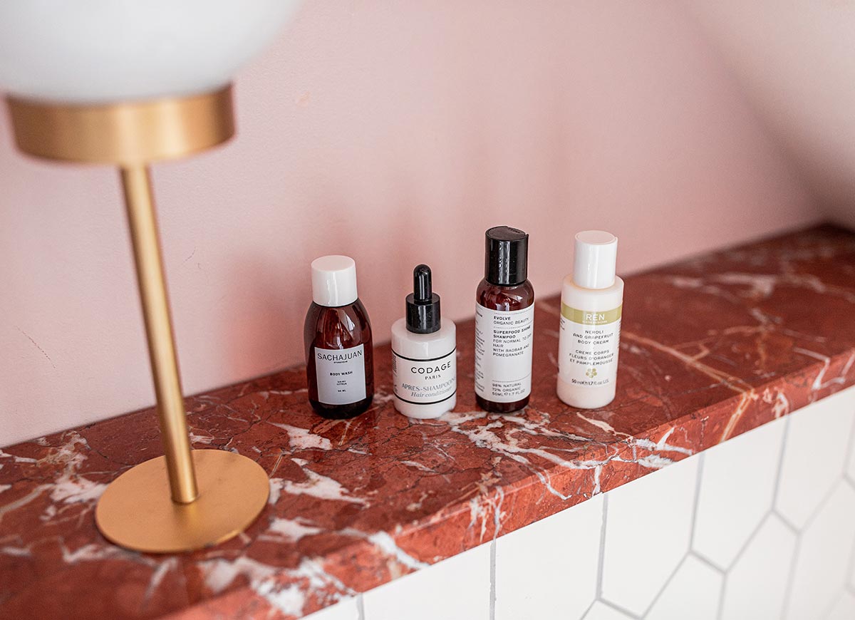 Close up of luxury bathroom amenities arranged on a red marble shelf against a soft pink wall. Bottles of body wash, shampoo, conditioner, and skincare products are neatly displayed beside a modern brass lamp, creating an elegant boutique hotel bathroom setting.