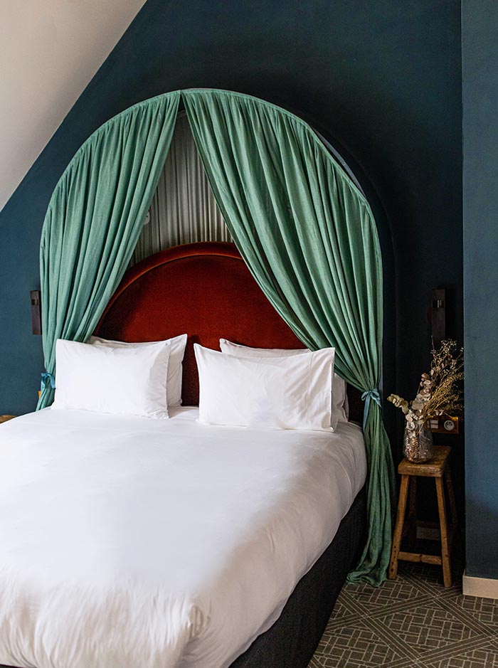 Canopy style hotel bed with crisp white linens framed by soft green curtains and a curved red upholstered headboard. Deep blue walls and warm accent decor create a cozy and sophisticated bedroom with a boutique hotel feel.