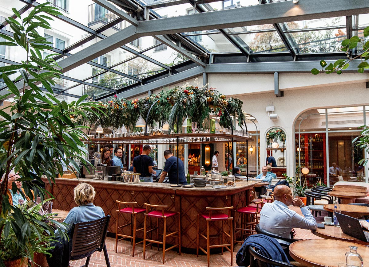 Bright hotel atrium restaurant with a glass ceiling, lush greenery, and a central bar surrounded by guests dining and socializing. Natural light fills the indoor courtyard space, creating a vibrant and inviting atmosphere that blends garden and café elements.