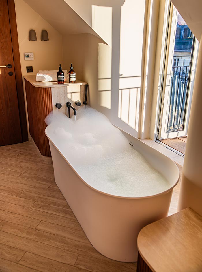 Freestanding bathtub filled with bubbles beside tall windows that open to a small balcony, with warm sunlight streaming across the room. Wood accents, soft neutral tones, and luxury bath amenities create a bright and relaxing spa-like retreat.