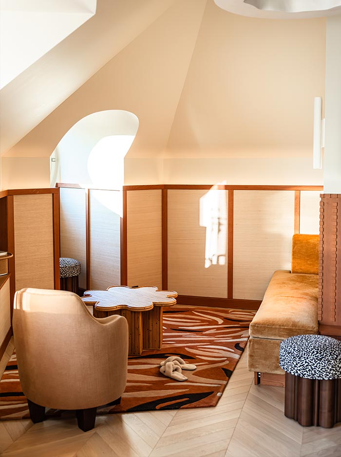 Cozy attic style sitting area with sloped ceilings, warm wood paneling, and soft natural light streaming through an arched window. A patterned rug, upholstered seating, and sculptural coffee table create an intimate and inviting space for relaxing within the hotel suite.