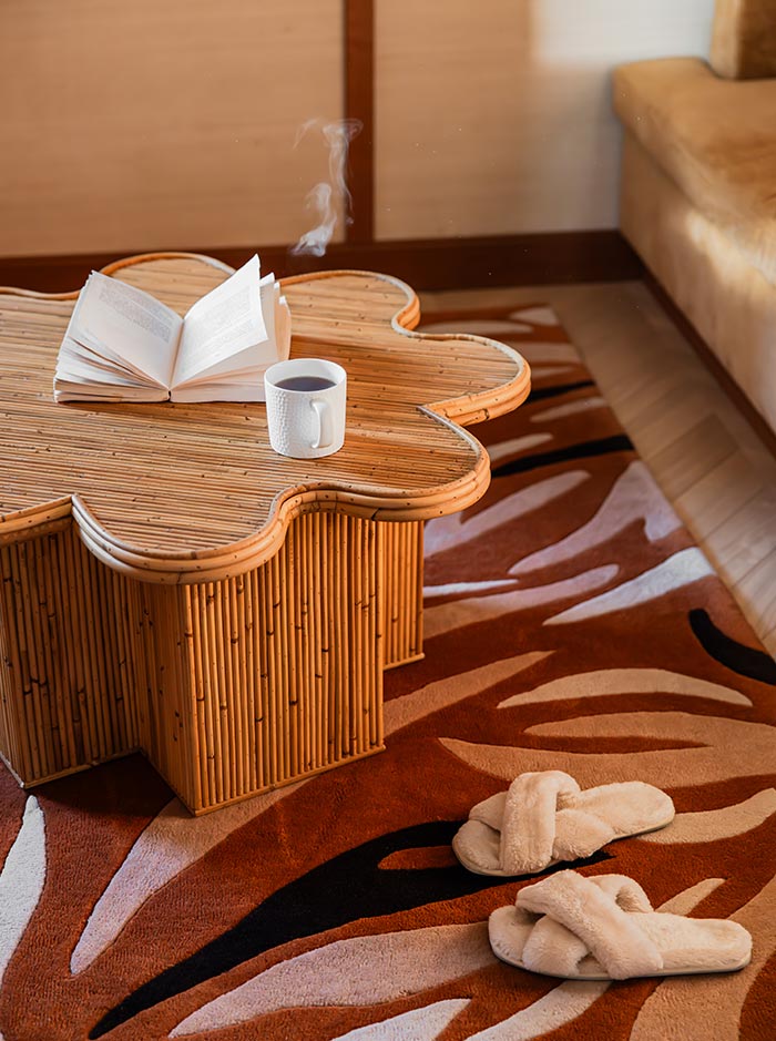 Open book and a steaming cup of coffee rest on a sculptural bamboo coffee table, creating a cozy reading nook within a hotel suite. Plush slippers lie on a patterned rug nearby, adding to the warm and relaxing atmosphere.