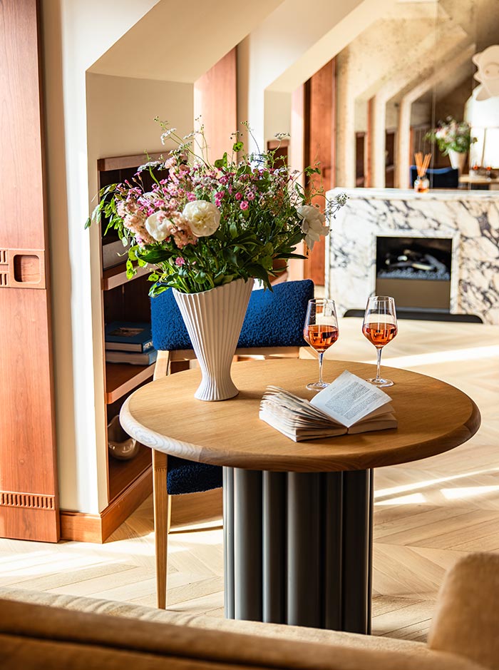 Sunlit hotel suite sitting area with a round wooden table holding fresh flowers, two glasses of rosé, and an open book. Warm wood accents, a marble fireplace, and soft natural light create an elegant and relaxing atmosphere.