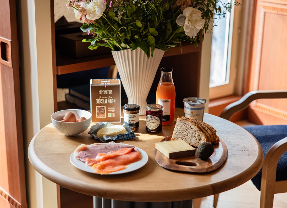 Round dining table set with breakfast ingredients including eggs, smoked salmon, bread, cheese, yogurt, juice, and preserves in a stylish hotel suite. A large bouquet of fresh flowers serves as the centerpiece, creating a warm and inviting atmosphere for an in room meal.