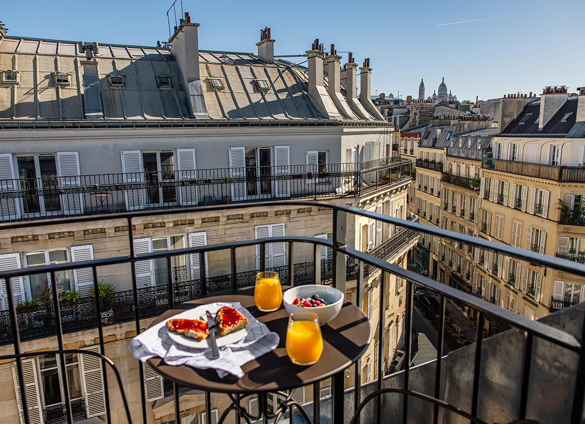 Small balcony breakfast setup with toast, fresh berries, and glasses of orange juice overlooking classic Parisian rooftops and elegant apartment buildings. Sunlight illuminates the cityscape while a quiet street view and distant landmarks create an authentic Paris neighborhood atmosphere.