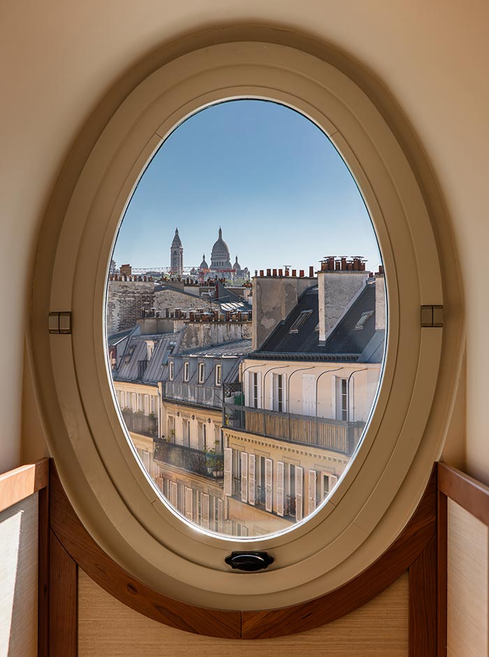 View of Paris rooftops framed through an oval window, with the dome and bell tower of Sacré Cœur visible in the distance beneath a clear blue sky. The unique window design creates a charming architectural frame for the classic cityscape beyond.