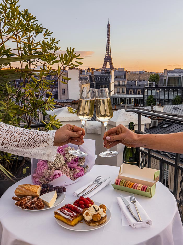 Two people toast with glasses of champagne on a rooftop terrace overlooking the Paris skyline, with the Eiffel Tower centered in the background at sunset. A table set with pastries, macarons, charcuterie, cheese, grapes, and a bouquet of flowers creates a romantic rooftop dining experience with iconic city views.