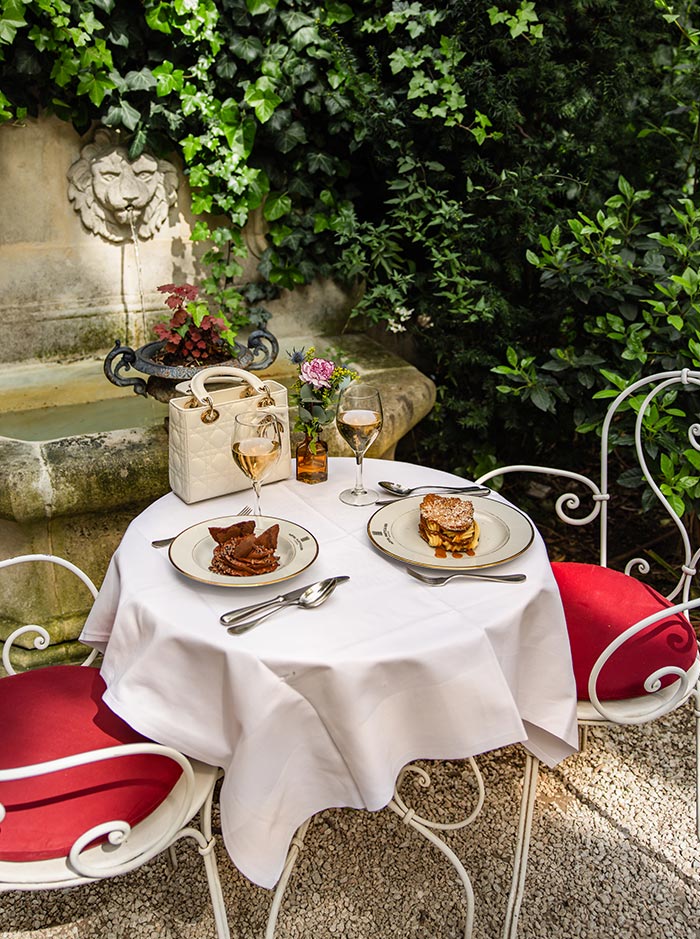 Romantic garden dining table with plated desserts and rosé wine beside a stone fountain, surrounded by lush greenery and elegant wrought iron chairs.