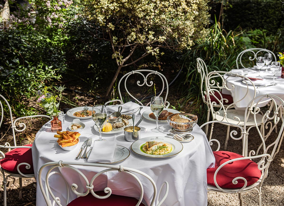 Outdoor garden dining table set for lunch with seafood dishes, white wine, fresh bread, and elegant place settings surrounded by lush greenery and wrought iron chairs.