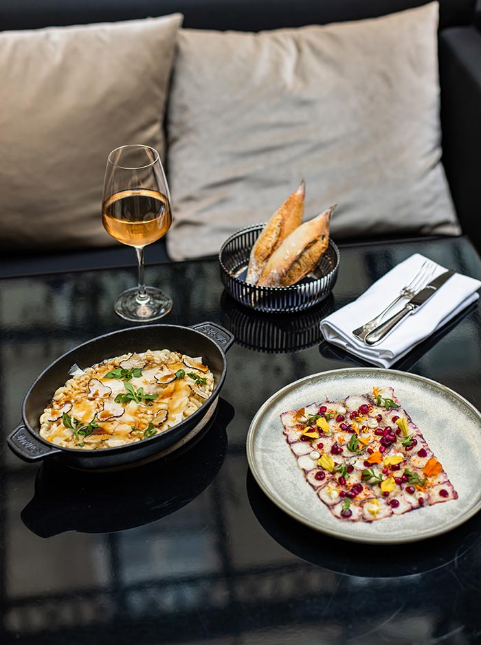 Restaurant table set with gourmet dishes, including a creamy truffle pasta and a colorful seafood carpaccio garnished with herbs and pomegranate seeds. A glass of white wine and basket of bread complete the elegant dining experience in a stylish lounge setting.