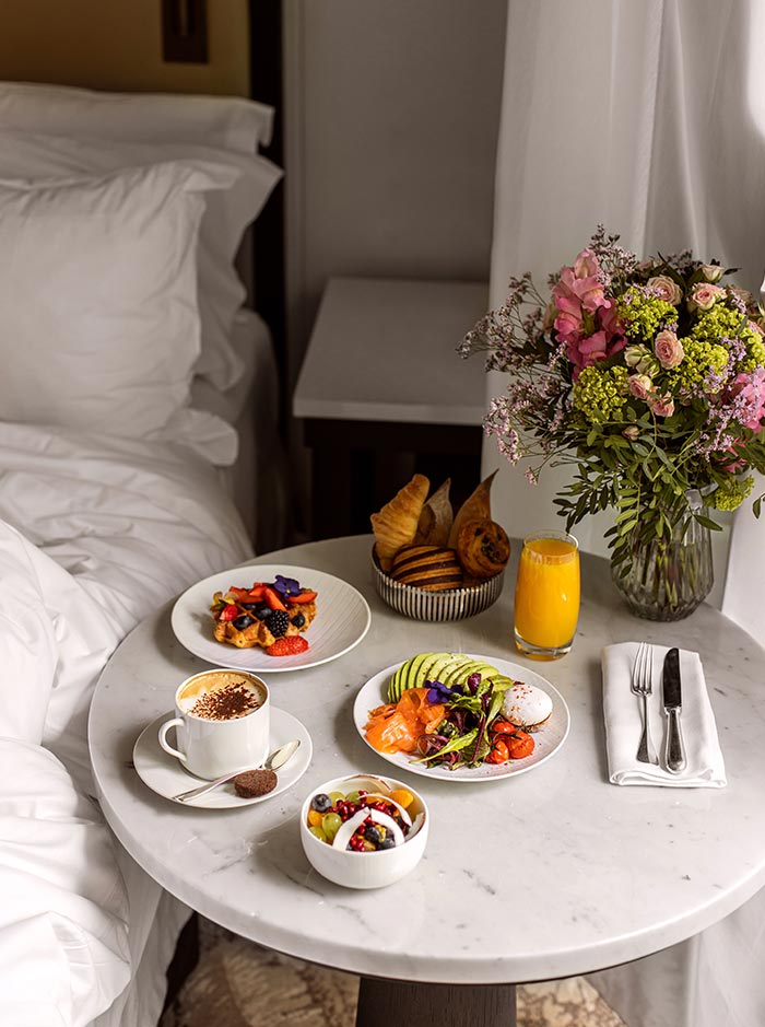 Breakfast spread on a marble table beside a hotel bed, featuring coffee, fresh fruit, pastries, orange juice, waffles with berries, and a smoked salmon and avocado dish. A bouquet of pink flowers adds color to the elegant in room dining setup, creating a luxurious and inviting morning atmosphere.