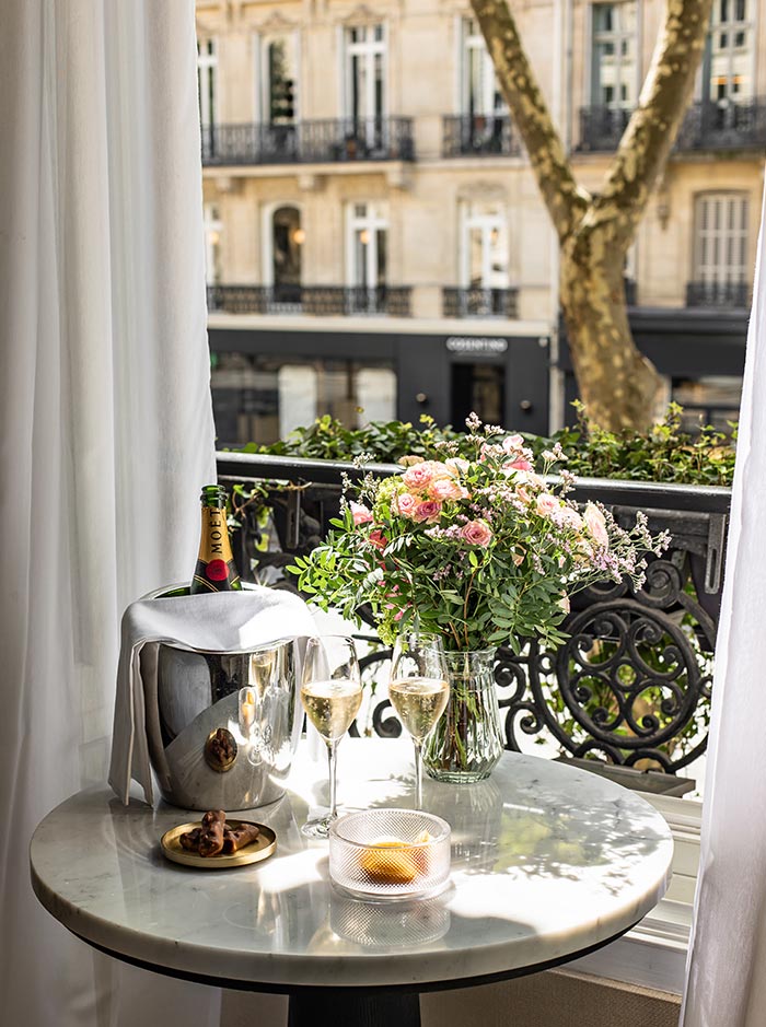 Round marble table beside an open balcony window set with chilled champagne, two glasses, and a bouquet of pink flowers. Sheer curtains frame the view of classic Parisian architecture and tree lined streets, creating an elegant and romantic hotel suite setting.