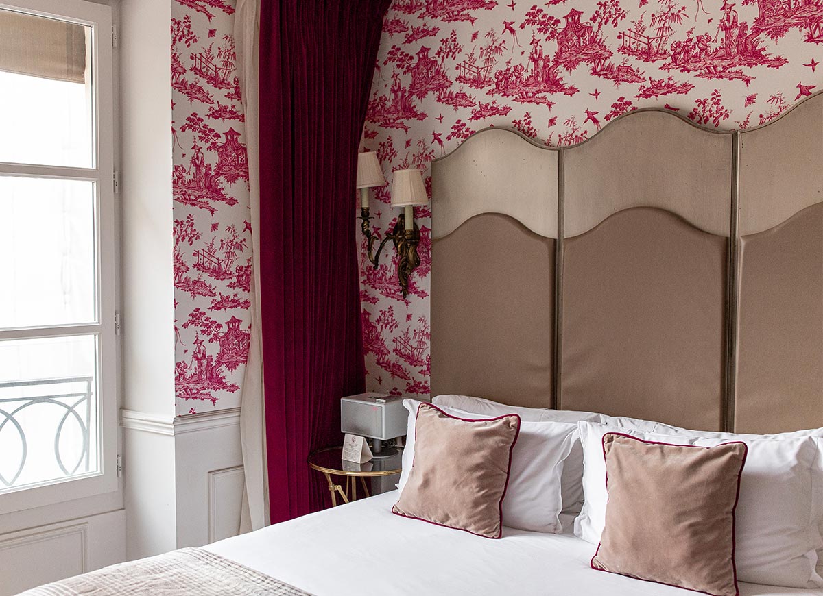 Elegant hotel bedroom with a tall upholstered headboard, crisp white bedding, and blush velvet accent pillows. Deep red curtains and ornate pink patterned wallpaper create a romantic boutique hotel atmosphere, while natural light streams in through the nearby window.