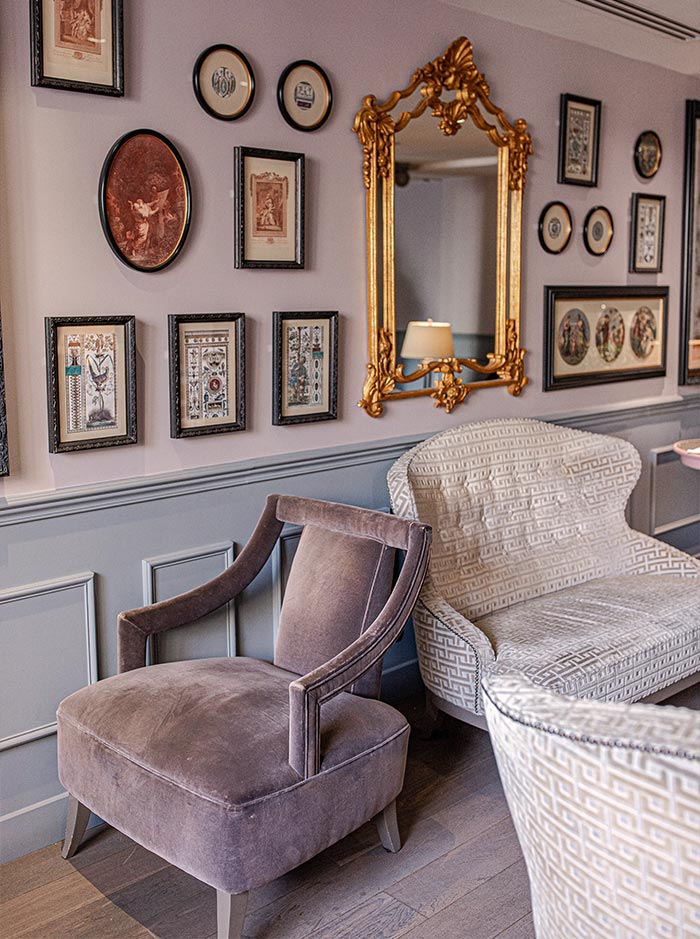 Elegant sitting area with upholstered armchairs in soft neutral tones, set against a gallery wall of framed artwork and vintage illustrations. An ornate gold mirror serves as the focal point, adding classic charm and sophistication to the refined interior.