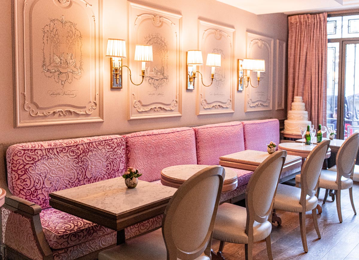Elegant café seating area with plush pink banquette seating, marble topped tables, and classic upholstered chairs arranged beneath ornate wall panels and warm sconces. Soft pastel tones, decorative detailing, and natural light from the windows create a refined and inviting Parisian tea room atmosphere.