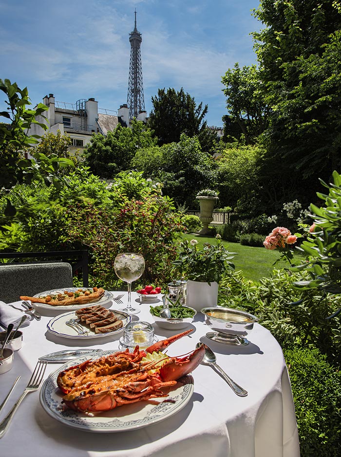 White tablecloth dining table set with grilled lobster, seafood dishes, and a glass of wine in a lush garden restaurant. The Eiffel Tower rises above the trees and Paris rooftops in the background, creating an elegant outdoor dining scene on a sunny day.