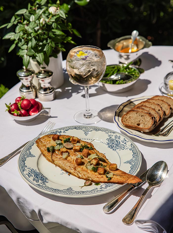 Whole grilled fish topped with herbs, capers, and golden croutons served on a patterned plate at an outdoor garden restaurant table. Fresh bread, radishes, green vegetables, and a large glass of sparkling wine complete the elegant al fresco dining setting.