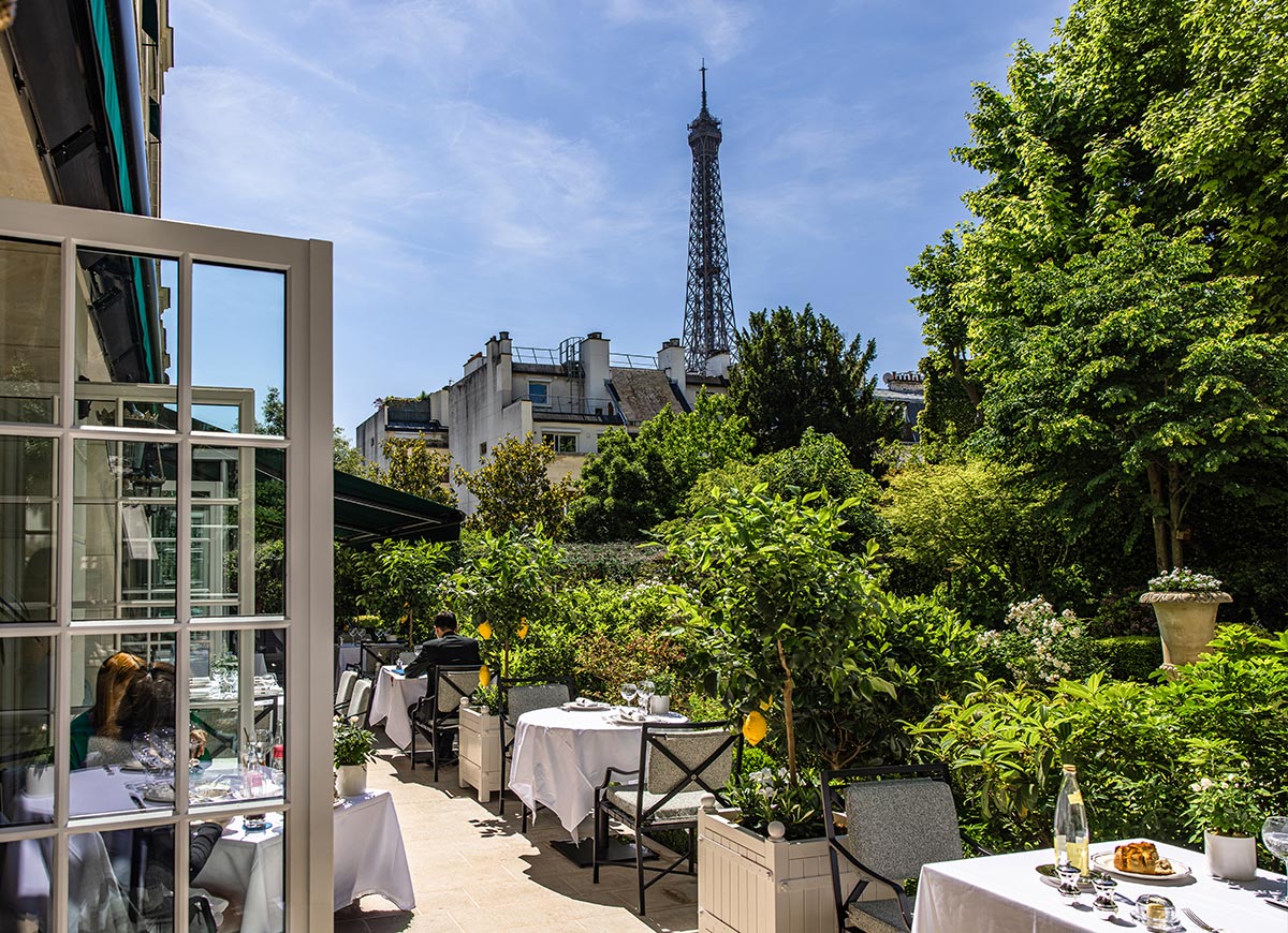 Outdoor garden restaurant terrace with white tablecloth dining tables surrounded by lush greenery and flowering plants. The Eiffel Tower rises above the trees and Paris rooftops in the background, creating a scenic setting for al fresco dining on a sunny day.