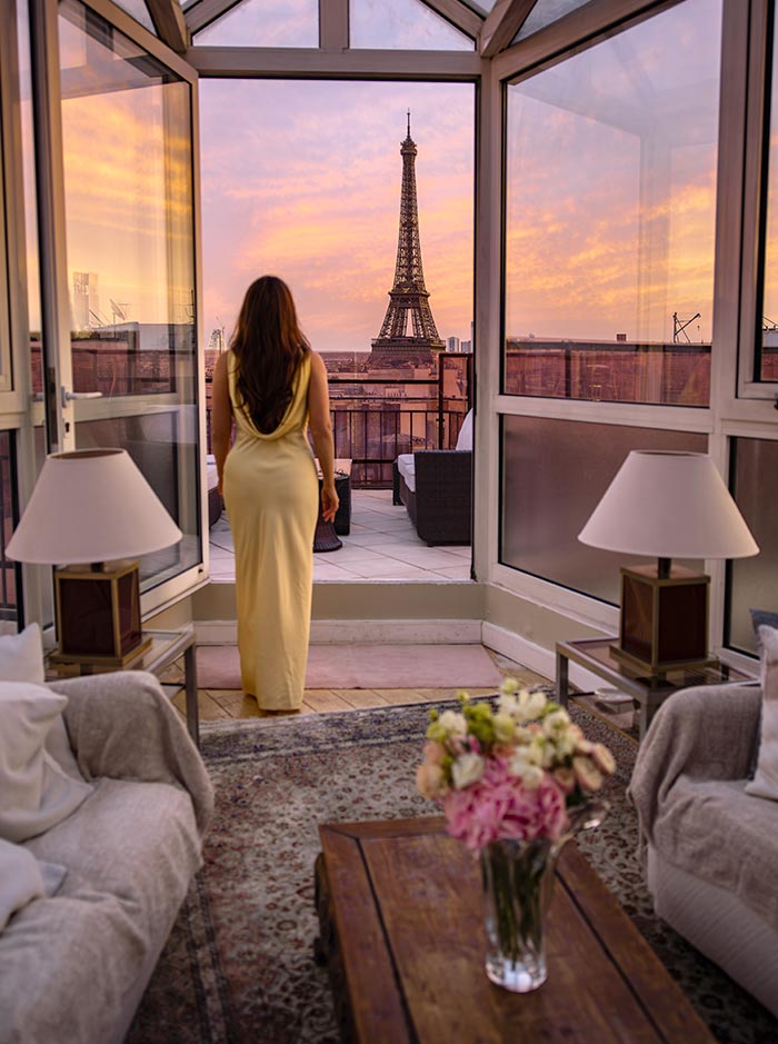 Person standing in a luxury hotel suite looking out toward the Eiffel Tower through open glass doors at sunset. Warm pink and orange skies frame the iconic landmark, while elegant furnishings and a bouquet of flowers create a romantic Parisian retreat.