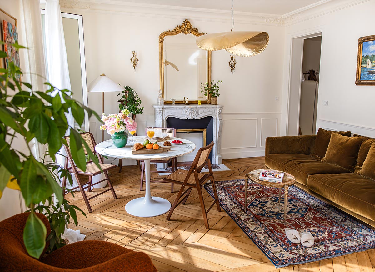 Elegant hotel suite living room with a velvet sofa, marble fireplace, and herringbone wood floors illuminated by natural sunlight. A round dining table set with pastries, fruit, flowers, and orange juice creates a welcoming atmosphere, while ornate details and classic furnishings add Parisian charm.