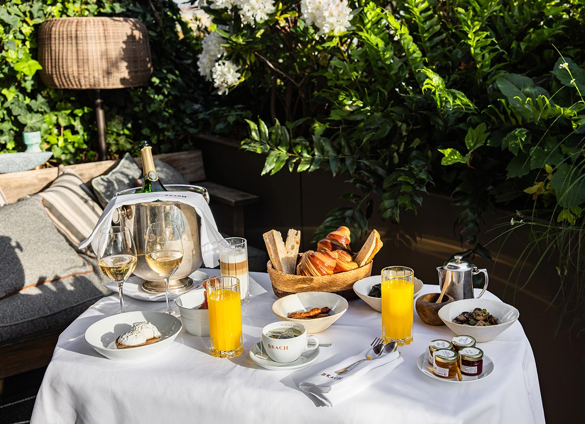 Breakfast table set in a lush garden terrace with croissants, coffee, fresh juice, preserves, and a chilled bottle of champagne. Surrounded by dense greenery and white flowers, the outdoor dining space creates an intimate and luxurious morning setting.