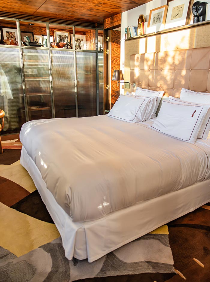 Cozy hotel bedroom with a large white bed, upholstered headboard, and warm wood paneled walls illuminated by natural sunlight. Framed artwork, books, and glass front wardrobes add character to the stylish and inviting guest room.