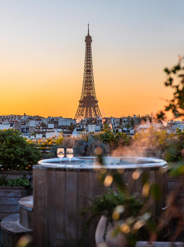 Steaming rooftop hot tubs overlook the Paris skyline at sunset, with the Eiffel Tower centered against a glowing orange sky. Wine glasses rest beside the tubs as soft evening light creates a romantic and luxurious atmosphere.