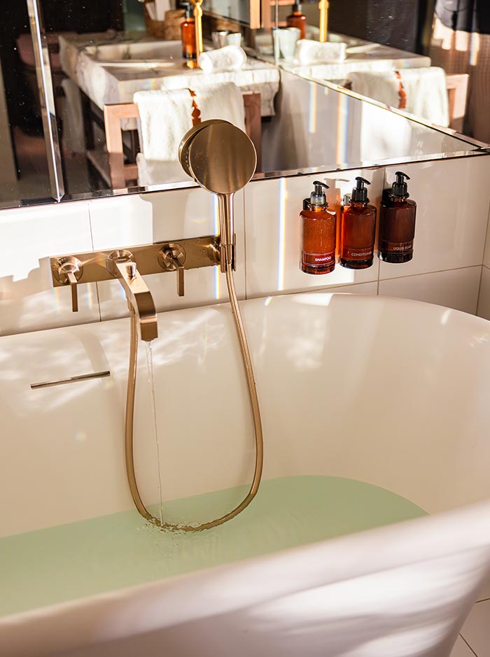 Freestanding bathtub filling with water beneath a large mirror, with brushed gold fixtures and amber bath amenities neatly arranged along the wall. Warm sunlight reflects across the bathroom, creating a calm and luxurious spa-like atmosphere.