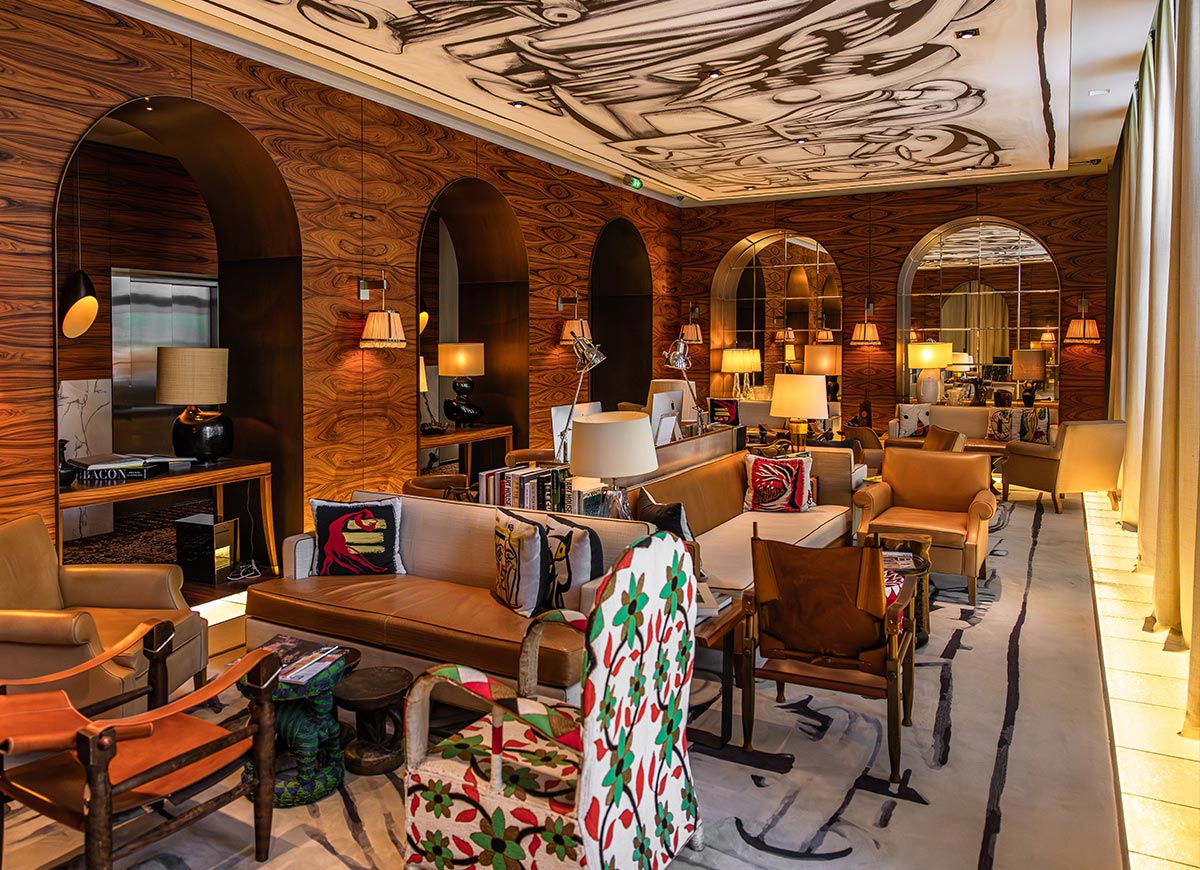 Warmly lit hotel lounge with rich wood paneled walls, arched mirrors, and a mix of leather and patterned seating arranged throughout the space. Decorative lighting, curated books, and artistic ceiling details create an inviting and sophisticated atmosphere.