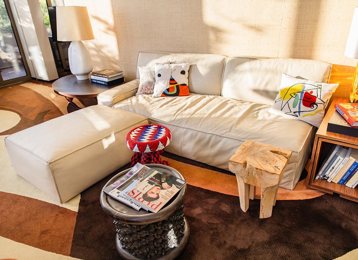 Sunlit hotel suite living area with a cream leather sofa, colorful decorative pillows, and sculptural wood furniture. Books, magazines, and modern artwork create a stylish and inviting space with a warm residential feel.