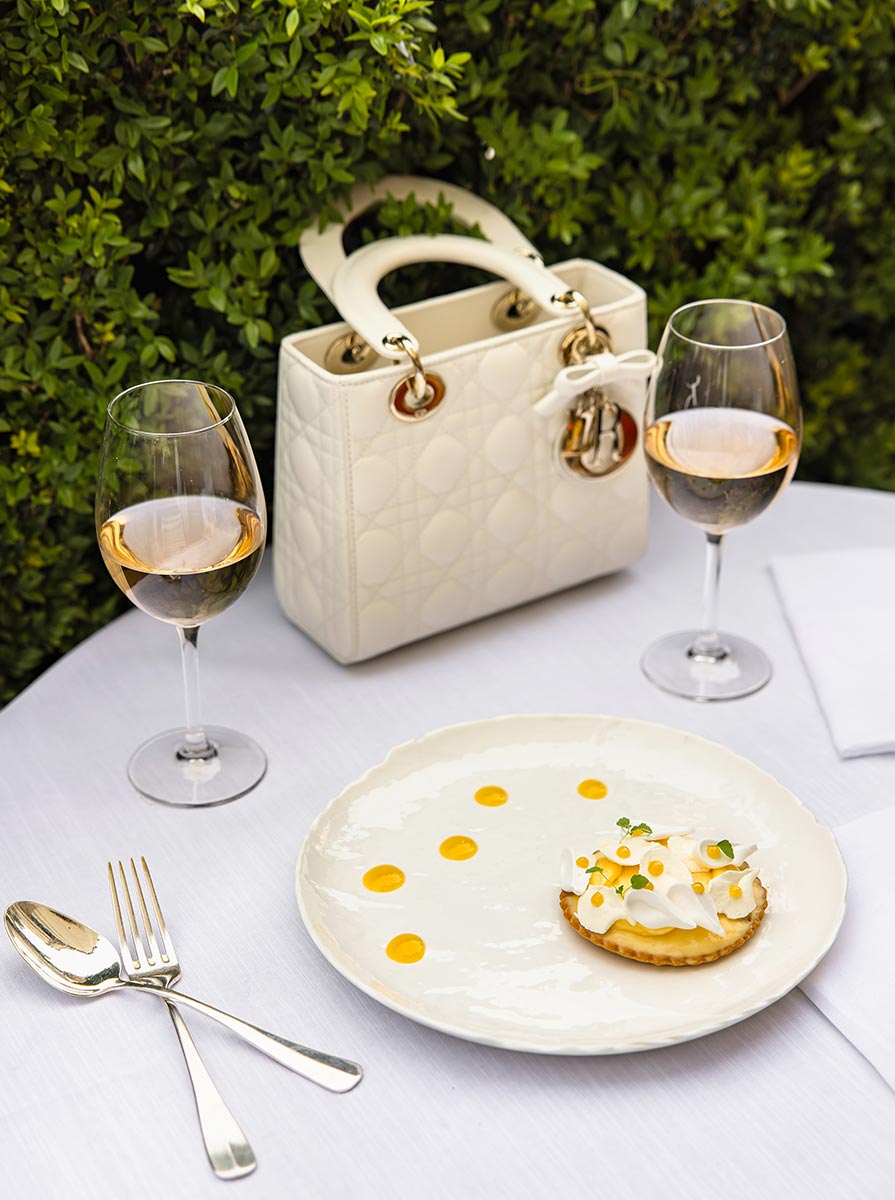 Elegant plated tart topped with delicate white cream curls and yellow sauce dots on a white tablecloth dining table. Two glasses of rosé wine and a white quilted handbag sit beside the dessert against a backdrop of lush green hedges at an outdoor Paris restaurant.