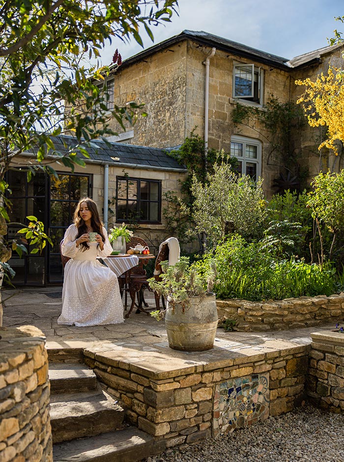 Woman in a flowing white dress seated at a small outdoor table in a sunlit stone courtyard garden. Rustic stone buildings, lush greenery, and potted plants surround the peaceful setting, creating a charming countryside atmosphere.
