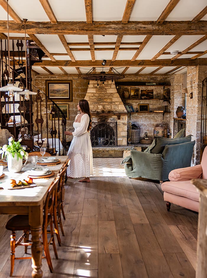 Woman in a white dress standing beside a stone fireplace inside a rustic countryside cottage with exposed wooden beams and warm natural light. A long wooden dining table, vintage spiral staircase, and cozy seating create an inviting farmhouse style interior.