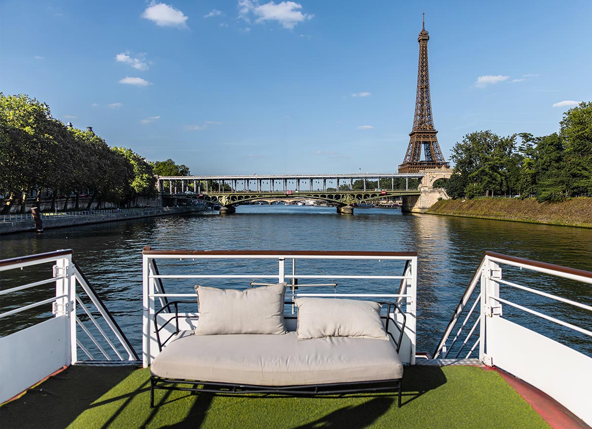 View from the deck of a river boat on the Seine with a cushioned lounge seat in the foreground and the Eiffel Tower rising beyond a bridge ahead. Trees line both banks of the river under a bright blue sky, highlighting a relaxed sightseeing experience in Paris
