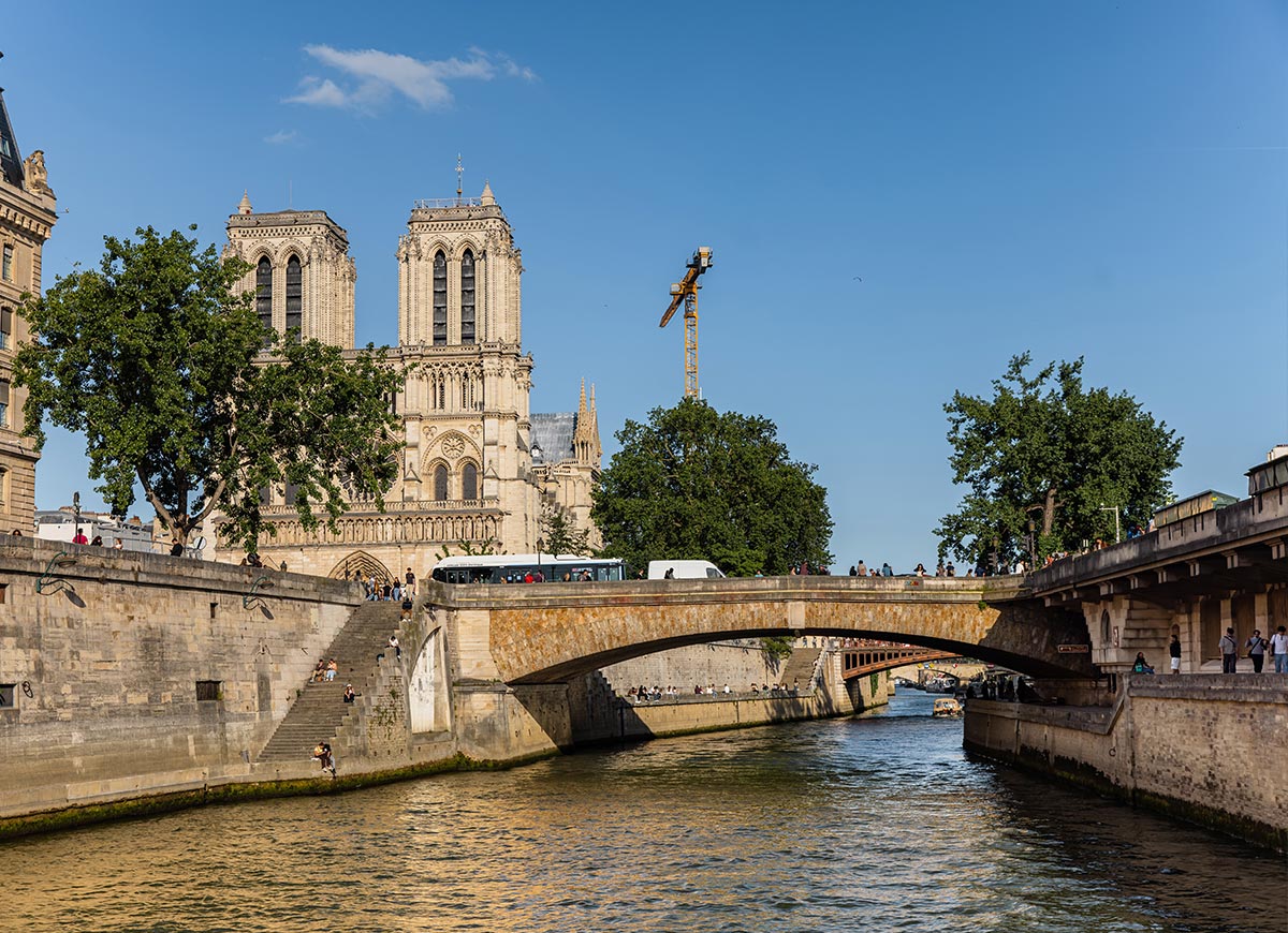 View of Notre Dame Cathedral and a stone bridge over the Seine River on a sunny day, with pedestrians and boats moving through the historic Paris cityscape.