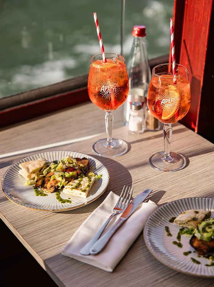 Two Aperol Spritz cocktails and plated sandwiches on a sunlit table beside a river view, creating a relaxed dining experience aboard a boat.