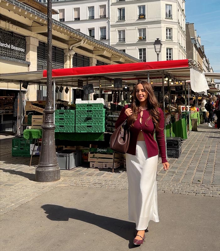 Woman walking through an outdoor market in Paris wearing a burgundy cardigan and white skirt, carrying a shoulder bag and smiling toward the camera. Market stalls with red awnings and fresh produce displays line the cobblestone street behind her.