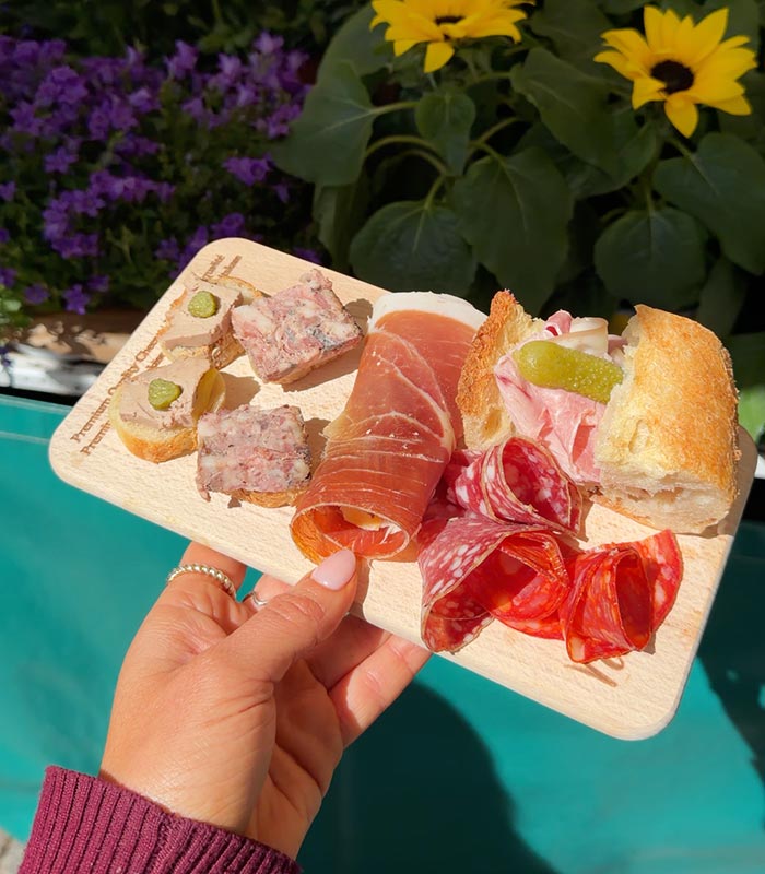 Hand holding a wooden charcuterie board topped with pâté crostini, cured meats, salami, and a ham sandwich with cornichons. Bright yellow sunflowers and purple flowers frame the scene beside a turquoise pool on a sunny day.