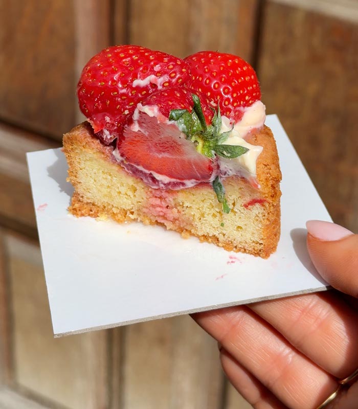 Slice of strawberry cake topped with fresh strawberries and cream, held by hand in bright sunlight. The golden sponge cake reveals a layer of strawberry filling, with a rustic wooden background softly blurred behind it.