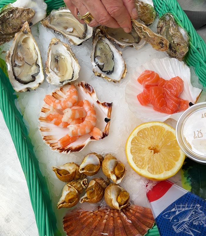 Seafood platter on crushed ice featuring freshly shucked oysters, shrimp, salmon sashimi, sea snails, and scallops. A hand opens an oyster beside a sliced lemon, creating a fresh market style seafood dining scene.