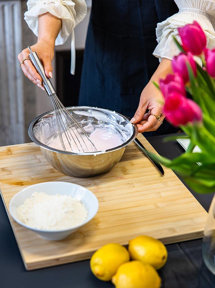 Close up of a baker whisking pink macaron batter in a mixing bowl beside lemons, flour, and fresh pink tulips on a kitchen countertop.