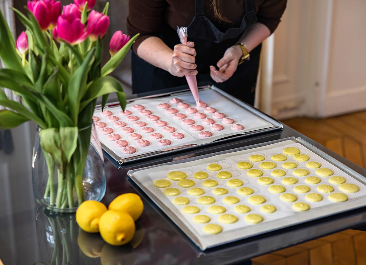Pastry chef piping pink and yellow macaron batter onto baking trays beside fresh tulips and lemons on a kitchen worktable.