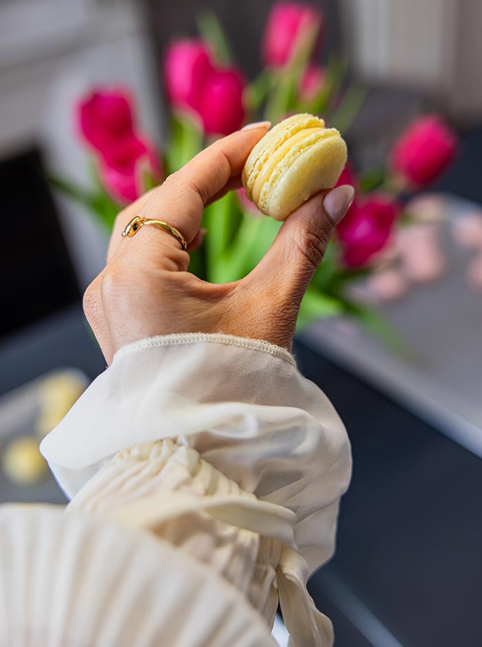 Hand holding a freshly made yellow macaron with pink tulips softly blurred in the background, highlighting the delicate texture and filling.