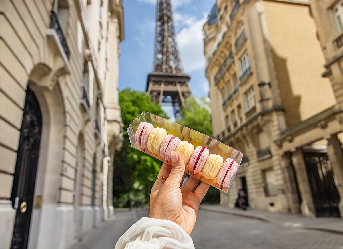 Hand holding a box of colorful macarons on a quiet Paris street with the Eiffel Tower rising in the background between historic buildings.