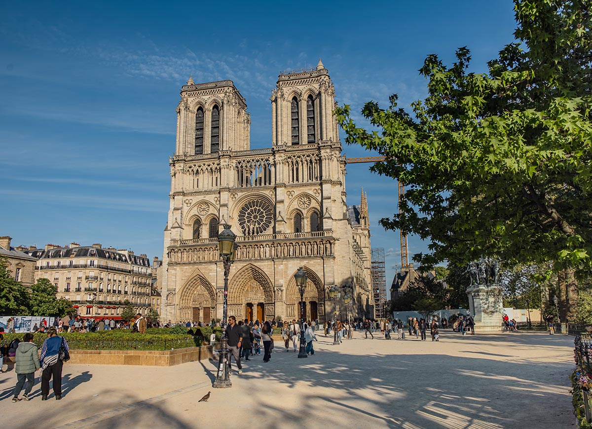 Wide view of Notre Dame Cathedral in Paris with visitors gathered in the plaza and trees framing the historic Gothic facade. The cathedral's twin towers and intricate stonework stand out against a clear blue sky.