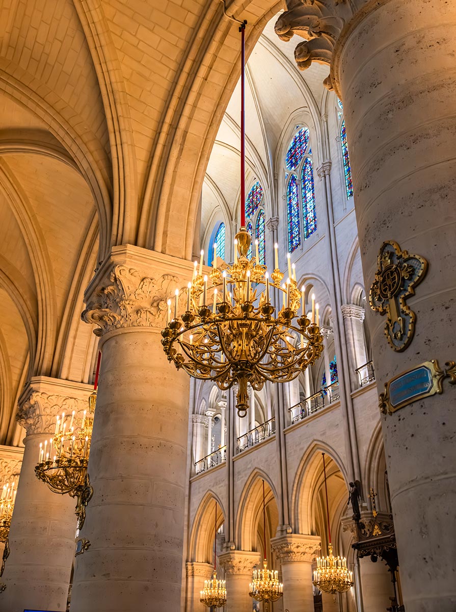 Close up of ornate gold chandeliers hanging from soaring stone arches inside Notre Dame Cathedral. Sunlight filters through colorful stained glass windows, highlighting the Gothic columns and intricate architectural details.