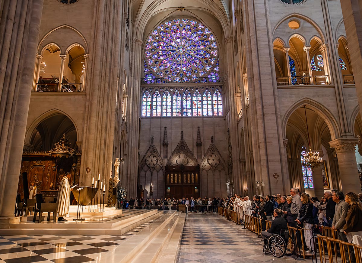 Congregation gathered inside Notre Dame Cathedral during a religious service, with clergy standing at the altar beneath towering Gothic arches. A large stained glass rose window fills the background, casting colorful light across the historic interior.