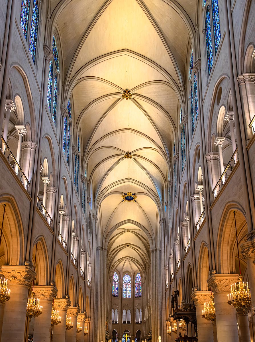 Interior view of Notre Dame Cathedral showcasing soaring vaulted ceilings, Gothic arches, and stained glass windows lining both sides of the nave. Warm chandeliers illuminate the grand stone columns and symmetrical architecture leading toward the altar.